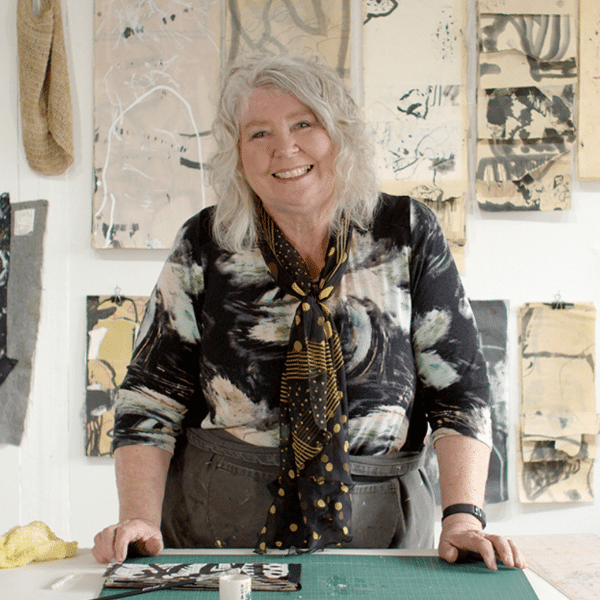 Image of artist Lorna Crane standing at her studio table,  smiling at the camera