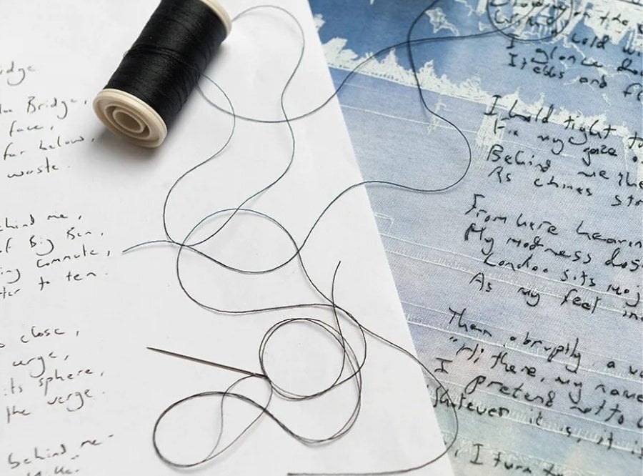 Handwriting on paper next to cloth, which the handwriting has been hand-stitched onto, with a spool of black thread and a needle.